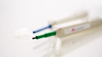 Close-up of fiber optic cleaning pens with visible tips, one green and one blue, placed on a reflective surface. The pen in focus is labeled with the Alcadon logo.