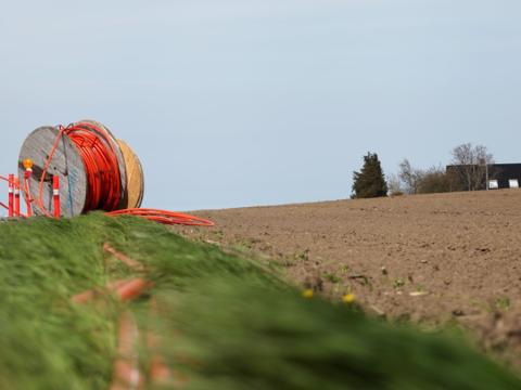 A large wooden spool of orange fiber optic cable placed alongside a rural field, with the cable partially unrolled and stretched along a grassy trench. A house is visible in the distance on the horizon.