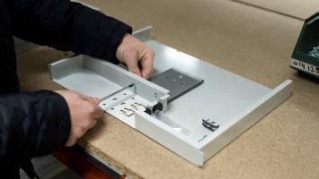 Person assembling a metal fiber distribution box on a workbench, aligning internal components and mounting brackets inside the enclosure.