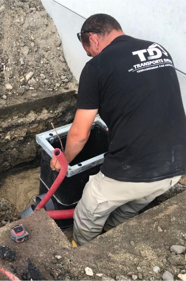 Worker installing a black underground utility chamber with red and grey conduits in a trench at a construction site, wearing a black T-shirt and beige pants