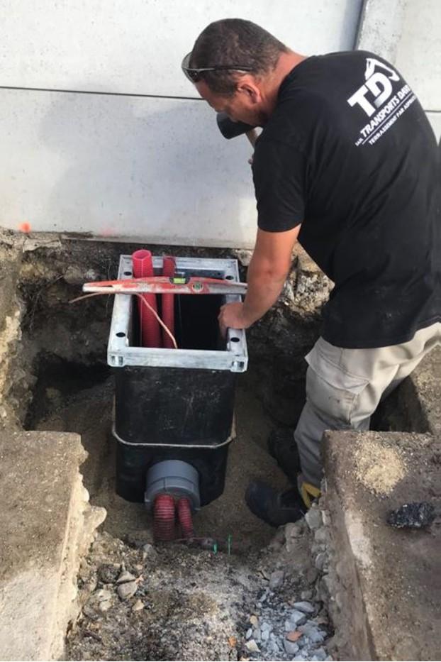 Technician aligning and installing an underground utility chamber with red conduits and a spirit level at a construction site, standing in a partially excavated trench