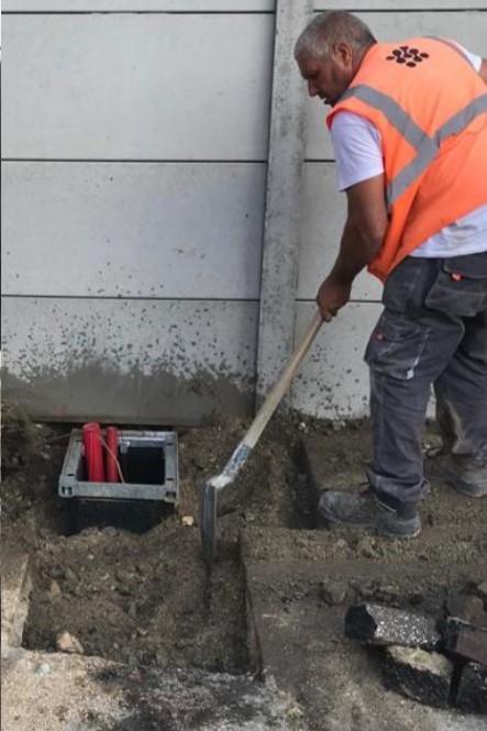 Construction worker in an orange safety vest using a shovel to backfill around an installed underground utility chamber with red conduits near a concrete wall