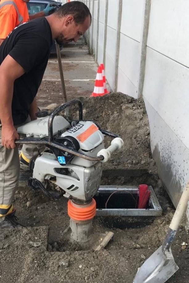 Worker operating a soil compactor to compress backfill around a newly installed underground utility chamber with red conduits next to a concrete wall