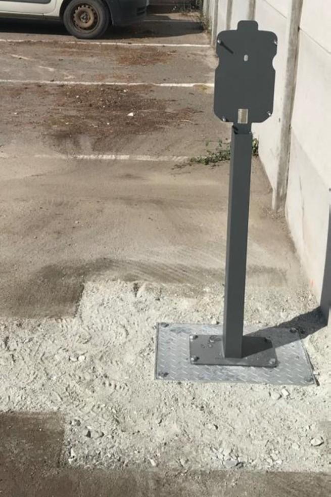 Installed outdoor power pedestal mounted on a metal base plate set in concrete, positioned along a wall on a paved area. A vehicle is parked in the background