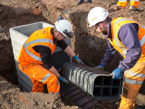 Two construction workers wearing safety gear and high-visibility clothing are installing a Cubis Systems plastic cable duct system into an underground chamber at a worksite, preparing for telecom or utility infrastructure.