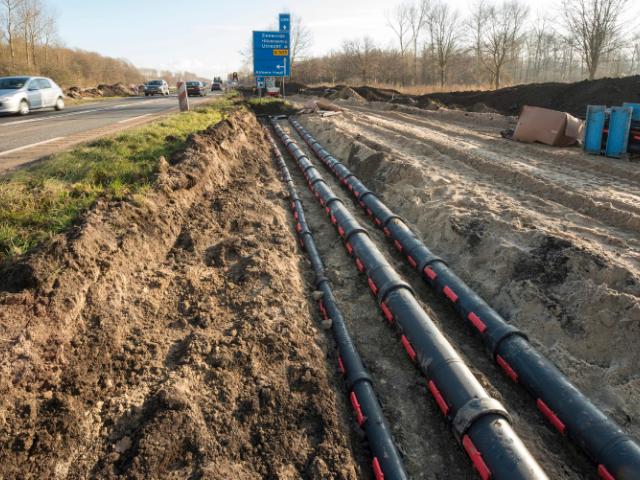 Underground cable protection system installation along a highway, showing multiple black conduits with red locking clips laid in a trench, with traffic passing by on the adjacent road.