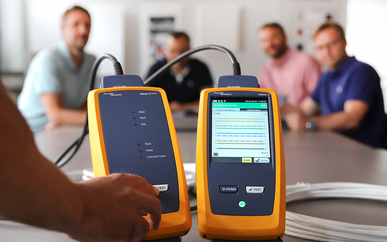 Close-up of a technician operating two yellow handheld network cable testers on a desk, with a group of four people blurred in the background discussing test results.