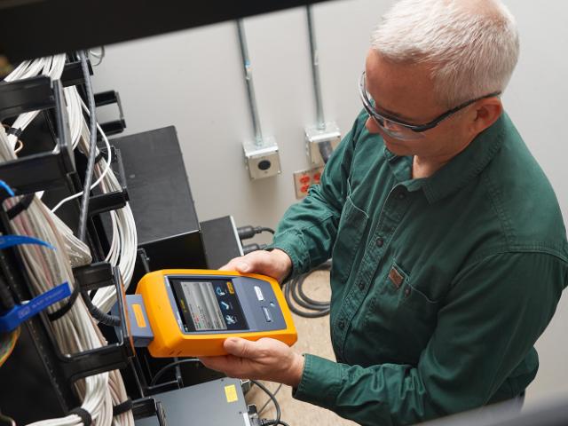Technician using a yellow handheld Fluke DSX network cable tester to check structured cabling connections in a server or telecom room, with organized white cables visible on a patch panel.
