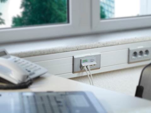 Office desk setup near a window, showing a network outlet with Ethernet cables plugged into a wall-mounted trunking system. A telephone and a laptop are partially visible in the foreground