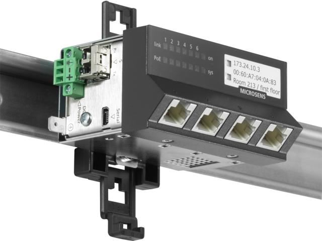 MICROSENS industrial Ethernet switch mounted on a DIN rail, featuring four RJ45 ports, fiber optic connection, and labeled IP configuration for network management.