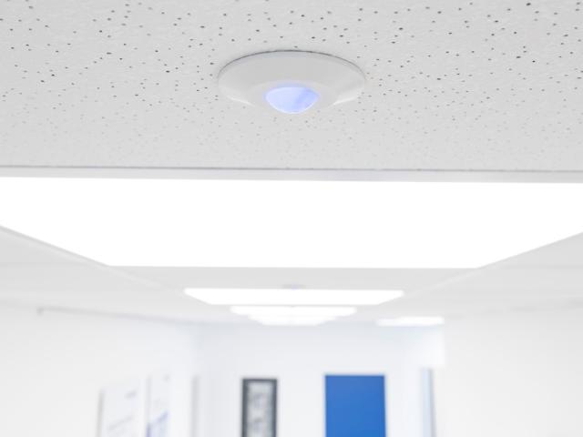 Close-up of a ceiling-mounted MICROSENS sensor or access point with a blue light indicator, integrated into a modern office ceiling with LED panel lighting.