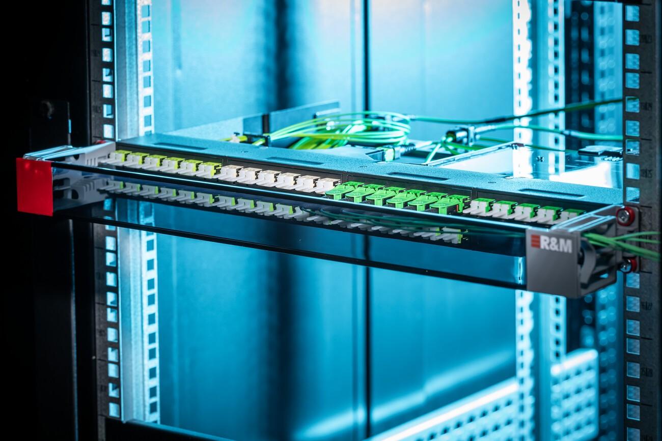 Fiber optic patch panel with multiple green connectors installed in a server rack, part of an R&M cabling system.