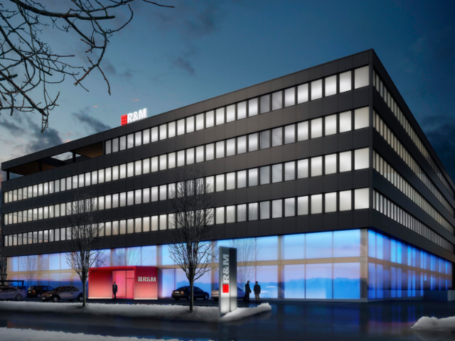 Modern office building at dusk with illuminated blue and red lighting on the ground floor. The building has five floors with uniform rectangular windows and bears the logo