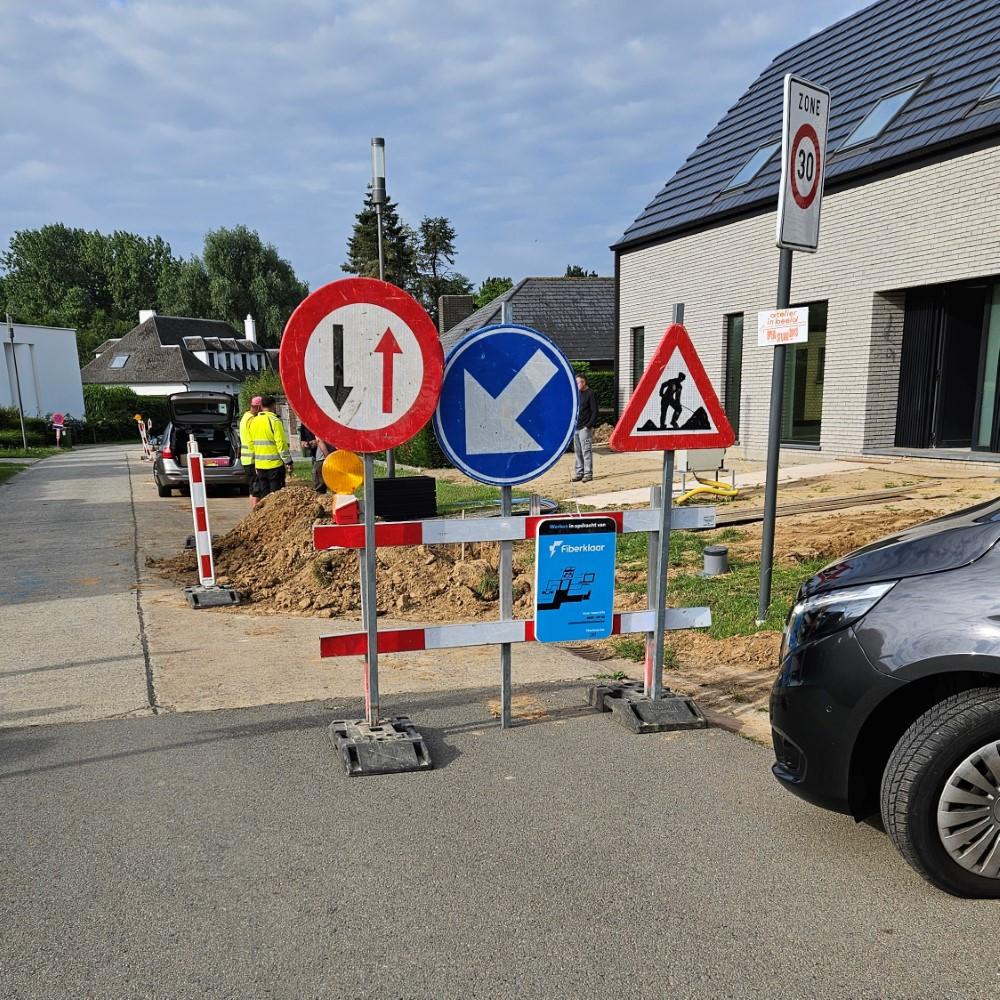 Roadwork in a residential area with multiple traffic signs in front of an excavation site, including signs for two-way traffic, keep left, roadwork, and a fiber installation warning. Two workers in yellow vests are seen near the dug-up area.