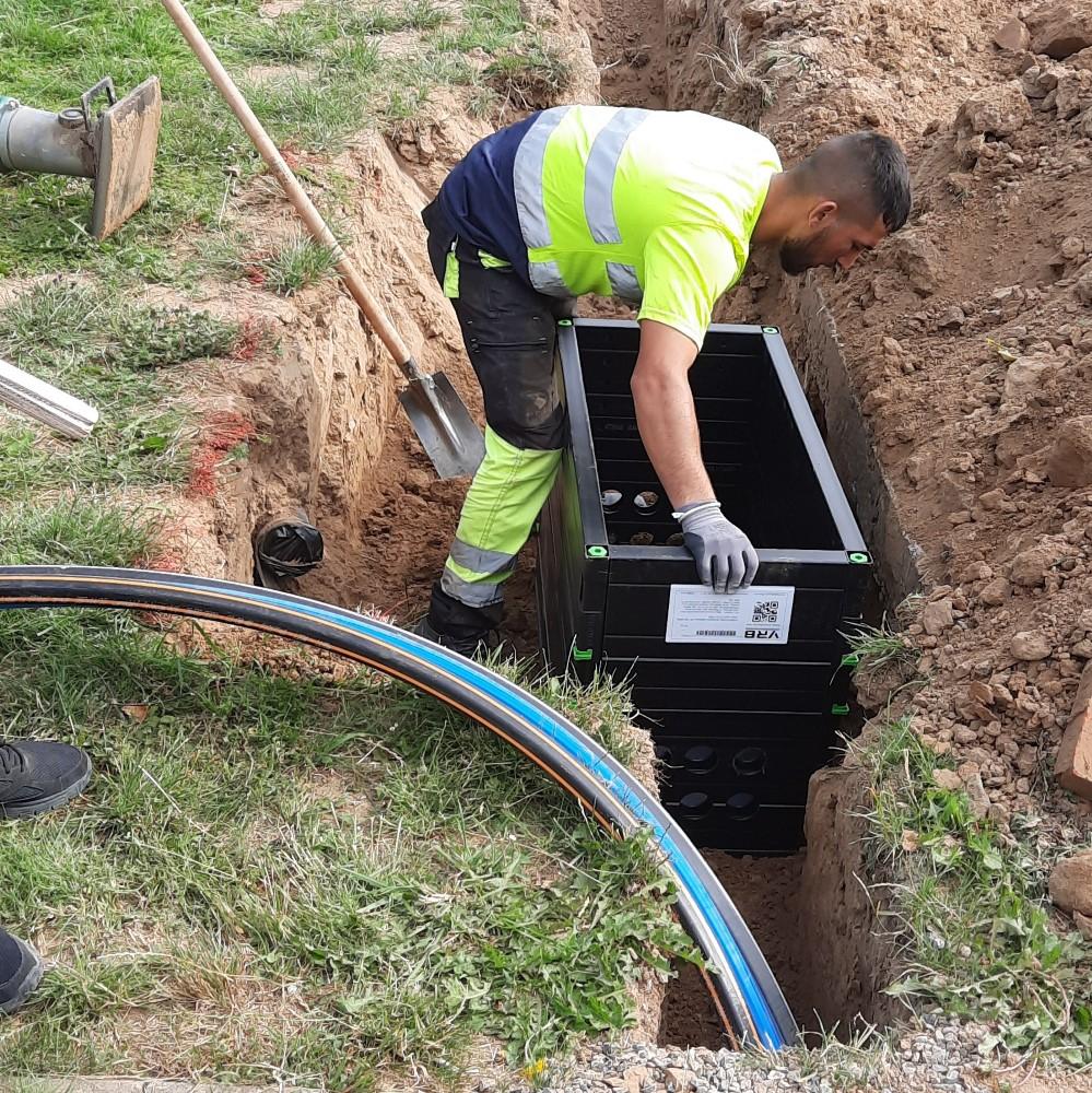 A worker in a high-visibility uniform is installing a black underground fiber optic distribution box in a trench. Several protective conduits for fiber cables are visible in the foreground, and a shovel lies nearby, indicating ongoing fiber installation.