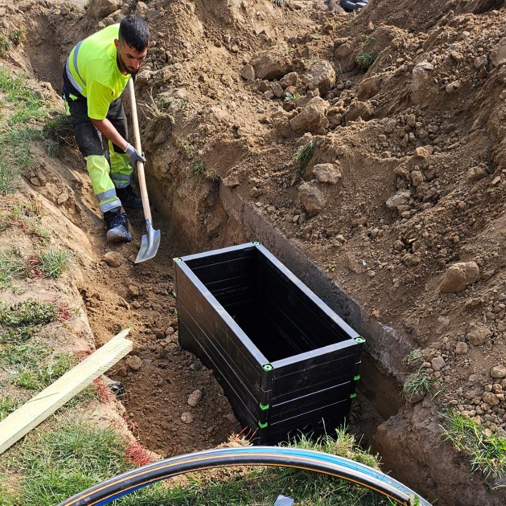 A worker in a neon yellow safety uniform is using a shovel to prepare the ground around a newly placed black underground fiber optic distribution box. The trench is deep, and several protective conduits for fiber cables are visible at the edge of the imag