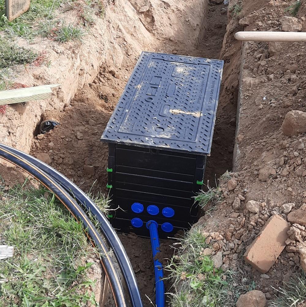 A newly installed underground fiber optic distribution box placed in a trench, with the lid securely mounted. Multiple blue microducts are connected to the box, and additional cable conduits are visible nearby, indicating active fiber installation work.