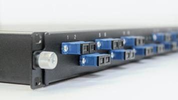 Close-up of a black fiber optic patch panel with multiple blue SC connectors inserted into numbered ports, set against a white background.